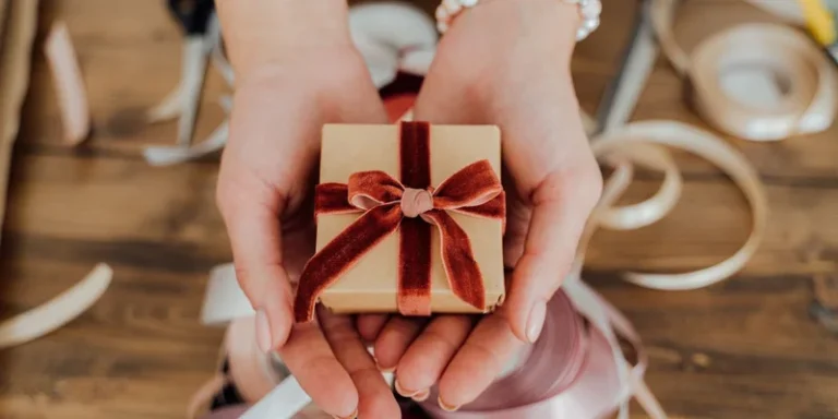 Hands presenting a small gift box wrapped with red velvet ribbon, surrounded by crafting materials